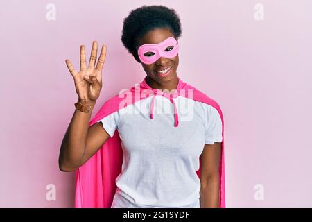 Junge afroamerikanische Mädchen mit Superhelden-Maske und Cape-Kostüm zeigt und zeigt mit den Fingern Nummer vier, während sie selbstbewusst und glücklich lächelt Stockfoto