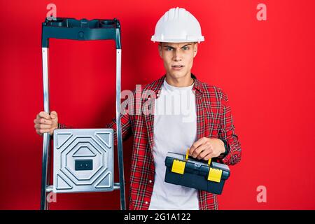 Junger hispanischer Mann, der einen Werkzeugkasten mit einem Hardhut an einer Bautreppe im Schockgesicht hält, skeptisch und sarkastisch aussagt und mit offenem Mund überrascht ist Stockfoto