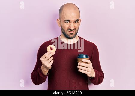 Junge Glatze Mann trinken Kaffee und essen Gebäck ahnungslos und verwirrt Ausdruck. Zweifel Konzept. Stockfoto