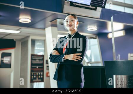 Porträt einer freundlichen weiblichen Bodenwärterin, die während einer Pandemie arbeitet. Frau mit Gesichtsschutz, die mit gekreuzten Armen am Flughafenterminal steht. Stockfoto