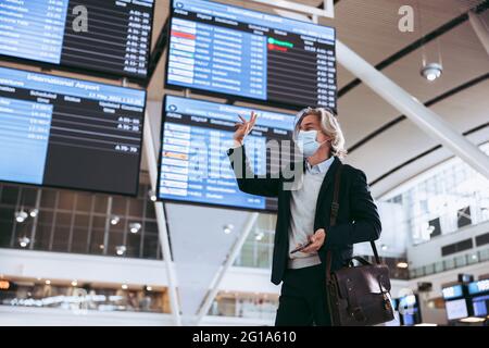 Mann mit Gesichtsmaske, der am Flughafenterminal steht und jemanden anwinkt. Geschäftsmann, der nach der Sperrung einer Pandemie reist. Stockfoto