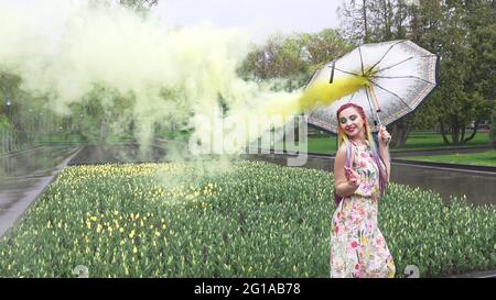 Entzückendes Mädchen in einem Kleid mit einem Blumendruck mit Regenbogen Zöpfen und Make-up. Tanzt mit einem Regenschirm, der sich in gelbem Rauch vor der Kulisse von FL Stockfoto