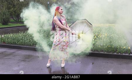 Entzückendes Mädchen in einem Kleid mit einem Blumendruck mit Regenbogen Zöpfen und Make-up. Tanzt mit einem Regenschirm, der sich in gelbem Rauch vor der Kulisse von FL Stockfoto