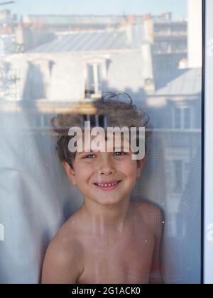 Ein süßer kleiner Junge steht im Zimmer am Fenster. Spiegelung im Fenster des gegenüberliegenden Hauses in Paris Stockfoto