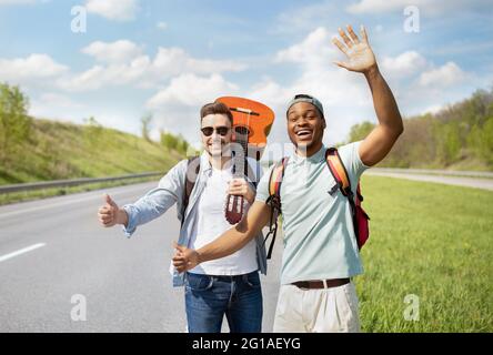 Zwei männliche Freunde mit Rucksäcken und Gitarre, die auf der Autobahn trampeln, Freifahrt machen, im Sommer zusammen reisen Stockfoto