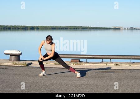 Eine junge Frau macht am frühen Morgen ein Warm-up auf dem Pier, vor dem Hintergrund des Flusses Stockfoto