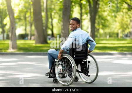 Fröhlicher behinderter schwarzer Mann im Rollstuhl, der Zeit im Stadtpark verbringt, in voller Länge Stockfoto