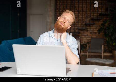 Müde rothaarige Geschäftsmann massiert Rücken des Halses, fühlt sich muskulös angespannt, junge bärtige kaukasische Freiberufler machen eine Pause von der Arbeit mit einem Laptop sitzen im Home Office Stockfoto