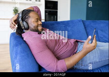 Ein glücklicher und entspannter afroamerikanischer Mann mit Kopfhörern verbringt seine Freizeit damit, Filme auf dem Smartphone zu sehen, ein schwarzer Mann in Kopfhörern, der sich auf dem Sofa niederlegt und mit dem Telefon telefoniert und Musik hört Stockfoto