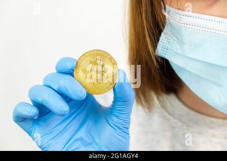 Nahaufnahme Frau Hand in Gummihandschuhe und schützende Gesichtsmaske hält Gold Bitcoin Münzen. Stockfoto