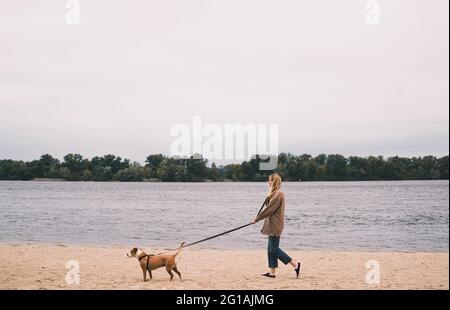 Frau geht bei düsterem Wetter am Ufer des Flusses am Sandstrand mit ihrem Hund spazieren Stockfoto