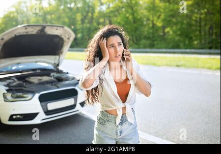 Tausendjährige kaukasische Frau mit Smartphone ruft den Abschleppdienst am Straßenrand an und steht in der Nähe ihres kaputten Fahrzeugs Stockfoto