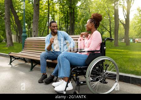 Positive behinderte schwarze Dame im Rollstuhl spricht mit ihrem Freund im Park, voller Länge Stockfoto