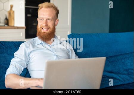 Fröhlicher charismatischer junger Rotschopf-Mann, der einen Laptop zum Studieren oder Online-Arbeiten verwendet, freiberuflicher Mann entwickelt ein neues Projekt, der aus der Ferne arbeitet, glücklicher bärtiger Hipster-Kerl schaut beiseite und träumt davon Stockfoto