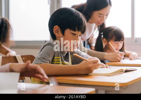 Asiatische Schullehrerin, die Schüler im Klassenzimmer unterstützt. Junge Frau, die in der Schule arbeitet und dem Jungen beim Schreiben, der Bildung, der Unterstützung und der Betreuung hilft. Stockfoto