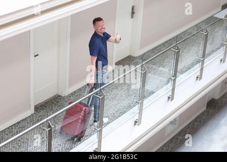Ein junger lächelnder Mann steht im Hotelkorridor und zeigt die Daumen nach oben Stockfoto