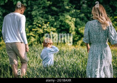 Glückliche Familie der jungen Bauern und ihr Sohn sind auf dem Feld mit üppigem grünen Gras zu Fuß. Rückansicht . Landwirtschaftskonzept Stockfoto
