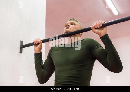 Der junge Sportmann in einem grünen Thermoanzug zieht sich an der horizontalen Stange im Fitnessstudio hoch. Stockfoto