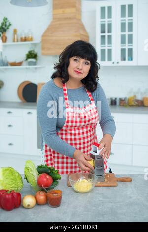 Frau in einer karierten Schürze reibt Käse auf einer Reibe in der Küche für Pizzabocken. Stockfoto