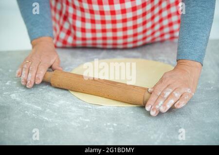 Die ziemlich reife Frau rollt den Teig mit einem Nudelholz in der Küche auf den Tisch. Oma Hausfrau. Stockfoto