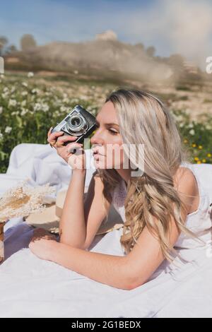 Positive Frau, die am Sommertag auf dem karierten Boden liegt und Fotos mit einer altmodischen Kamera auf dem Land fotografiert Stockfoto