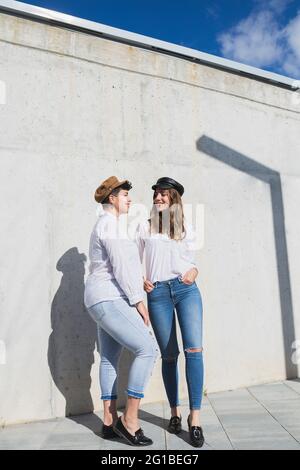 Ganzer Körper junger positiver Freundinnen in trendigen Outfits und Hüten, die sich an sonnigen Tagen auf dem Gehweg nahe der grauen Wand unter Blau anguckend sehen Stockfoto