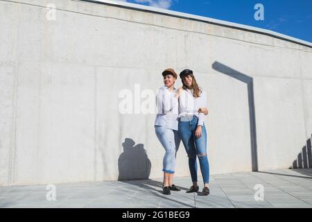 Voller Körper junger positiver Freundinnen in trendigen Outfits und Hüten, die an sonnigen Tagen unter blauem Himmel auf einem Gehweg in der Nähe einer grauen Wand stehen Stockfoto