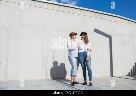 Ganzer Körper junger positiver Freundinnen in trendigen Outfits und Hüten, die sich an sonnigen Tagen auf dem Gehweg nahe der grauen Wand unter Blau anguckend sehen Stockfoto