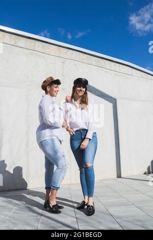 Voller Körper junger positiver Freundinnen in trendigen Outfits und Hüten, die an sonnigen Tagen unter blauem Himmel auf einem Gehweg in der Nähe einer grauen Wand stehen Stockfoto