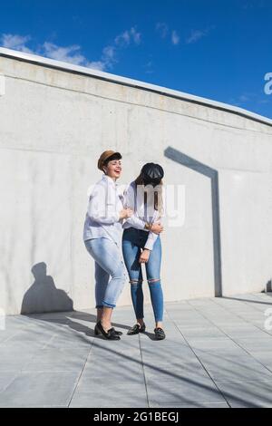 Voller Körper junger positiver Freundinnen in trendigen Outfits und Hüten, die an sonnigen Tagen unter blauem Himmel auf einem Gehweg in der Nähe einer grauen Wand stehen Stockfoto