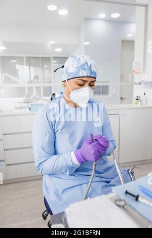 Ernsthafte ethnische Zahnärztin in Maske und medizinischer Uniform mit Handschuhen, die an Bohrmaschine in der Klinik arbeiten Stockfoto