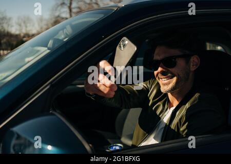 Glücklicher Mann in Sonnenbrillen, der Selfie auf dem Handy macht, während er am sonnigen Tag im Auto sitzt Stockfoto