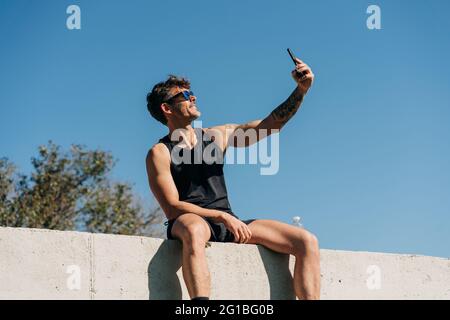 Setzen Sie den männlichen Athleten in Sportkleidung ein, indem Sie nach dem Training Selfie mit dem Mobiltelefon machen und sich auf dem Zaun ausruhen Stockfoto