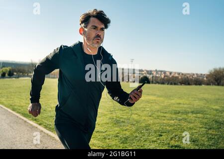 Der Jogger für Erwachsene in Sportbekleidung läuft auf dem Bürgersteig zwischen den Rasenflächen und freut sich während des Trainings in der Stadt auf die Zukunft Stockfoto