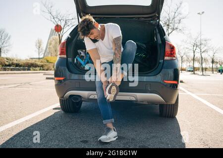 Tätowierte männliche Athletin, die Schuhe anlegt, während sie auf dem Handy gegen ein Auto mit offenem Kofferraum in der Stadt spricht Stockfoto