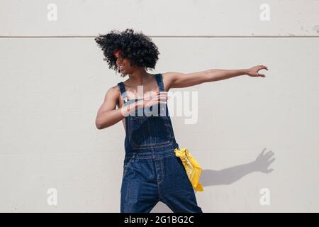 Fröhlicher afroamerikanischer Mann mit Afro-Frisur in Denim-Overalls, der auf Betonwänden tanzt und wegschaut Stockfoto