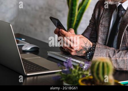 Schneiden Sie nicht erkennbare tätowierte männliche Führungskraft in karierter Jacke Textnachrichten auf dem Handy gegen Laptop während der Telearbeit Stockfoto