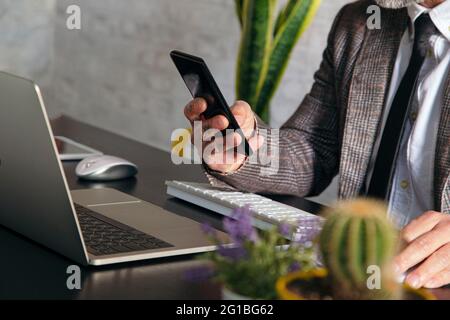 Schneiden Sie nicht erkennbare tätowierte männliche Führungskraft in karierter Jacke Textnachrichten auf dem Handy gegen Laptop während der Telearbeit Stockfoto
