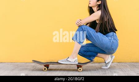 Seitenansicht der kurzgeschnittenen, nicht erkennbaren jungen verträumten Skateboarderin im lässigen Outfit, das sich auf dem Laufsteg hockt Stockfoto