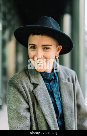 Fröhliche junge Transgender-Person in edlem Mantel und Hut, die bei Tageslicht auf die Kamera schaut Stockfoto