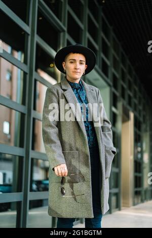 Junge Transgender-Person in edlem Mantel und Hut, die bei Tageslicht auf die Kamera schaut Stockfoto