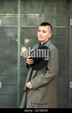 Junge Transgender-Person in edlem Mantel und Hut mit Mohawk, die bei Tageslicht auf die Kamera schaut Stockfoto