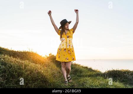 In voller Länge attraktives junges Weibchen in gelbem Sundress und Hut stehend mit Armen auf grüner Wiese in sonniger Landschaft angehoben Stockfoto