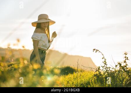 Attraktive, verträumte Hündin in Jeans und weißem Top und in angenehmen Gedanken wegschauen, während sie an sonnigen Tagen auf üppigem, grasbewachsenen Gelände steht Stockfoto