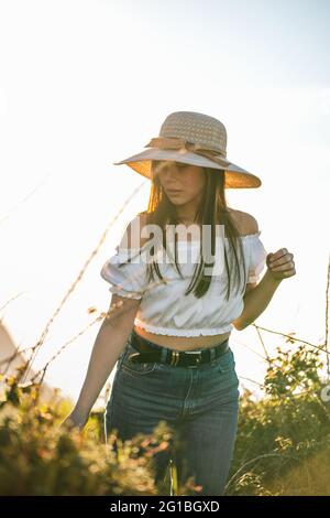 Attraktive, verträumte Hündin in Jeans und weißem Top und in angenehmen Gedanken wegschauen, während sie an sonnigen Tagen auf üppigem, grasbewachsenen Gelände steht Stockfoto