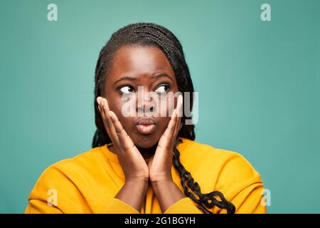 Frohes afroamerikanisches Weibchen in gelben Kleidern, die Lippen ausschüttet und wegschaut, während sie das Gesicht in den Händen vor blauem Hintergrund hält Stockfoto