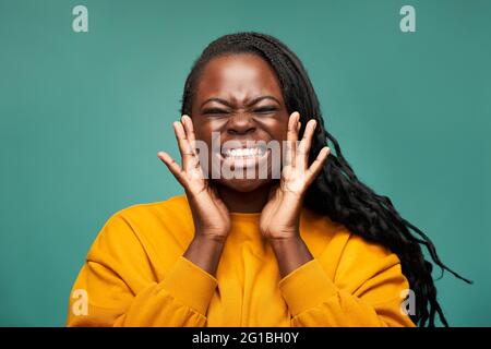 Frohes afroamerikanisches Weibchen in gelber Kleidung mit toothy Lächeln und geschlossenen Augen, das Gesicht in den Händen vor blauem Hintergrund haltend Stockfoto
