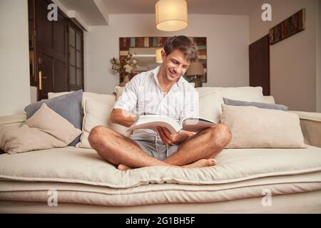 Lächelnder, erwachsener Mann in legerer Kleidung sitzt auf dem Sofa mit offenem Buch in den Händen in einem hellen Haus mit Kissen und Schrank Stockfoto