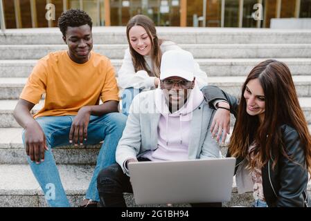 Verschiedene junge Freunde lächeln weit, während sie sich gemeinsam ein Video auf Netbook ansehen, während sie auf Betontreppen sitzen Stockfoto