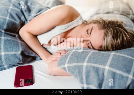 Junge Frau, die morgens auf einem weichen Kissen unter der Decke im Bett in der Nähe des Mobiltelefons schläft Stockfoto